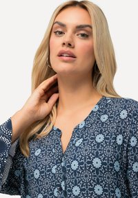 Blusa de estampado floral azul con cierre de botón. Presenta un escote en V y mangas acampanadas con un acento de puntos. Textura de tela suave.