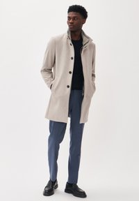 Abrigo de mezcla de lana gris claro con un cuello alto, botones negros, bolsillos, usado sobre un suéter negro y pantalones azules, combinado con zapatos negros.