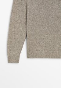 Ærme og nederste del af en lysebrun strikket sweater med ribbede manchetter og kant mod hvid baggrund.