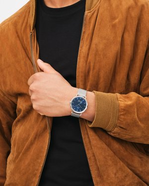 Montre analogique pour homme avec un cadran bleu, un bracelet en maille argentée et un boîtier rond argenté, portée à un poignet contre une veste en daim marron.