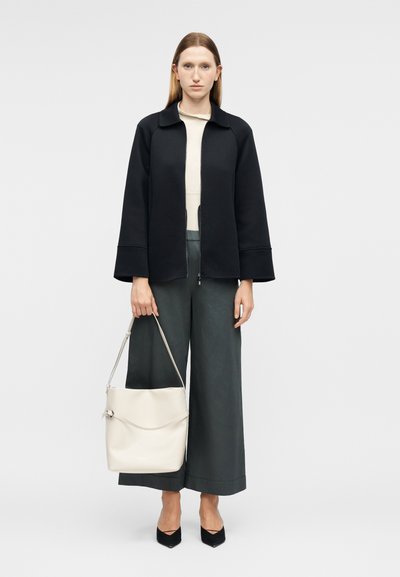 Femme aux cheveux raides portant une veste noire, un haut crème, un pantalon large gris, des talons noirs, tenant un grand sac d'épaule blanc.