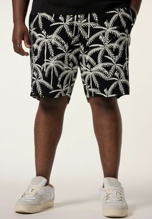 Mand iført sorte shorts med hvidt palmetræprint og hvide sneakers, stående med hænderne i lommerne.