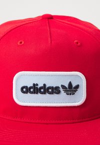 Rdeča kapa s ravnim robom. Vsebuje bel pravokotni vložek z logotipom "adidas" v črni vezenini in trefoil logotipom. Gladka struktura tkanine.