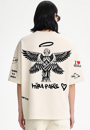 Persoon die een beige overhemd draagt met een zwarte engelengrafiek, halo, tekst ""Hira Paris"" en verschillende zwarte en rode ontwerpen op de rug en mouwen.