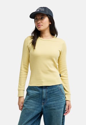 Femme portant une casquette de baseball noire, une chemise à manches longues jaune clair et un jean bleu, debout et regardant légèrement sur le côté.