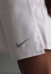 Ανοιχτό ροζ αθλητικά σορτς με γκρί λογότυπο Nike swoosh κοντά στο τελείωμα στο μηρό ενός ατόμου, με δίχτυ στο πλάι.