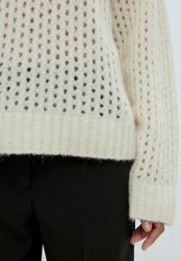 Strickpullover in Creme mit einem offenen Webmuster, das einen Farbverlauf von braun zu schwarz zeigt. Ribbelbündchen und Saum, weiche Textur.