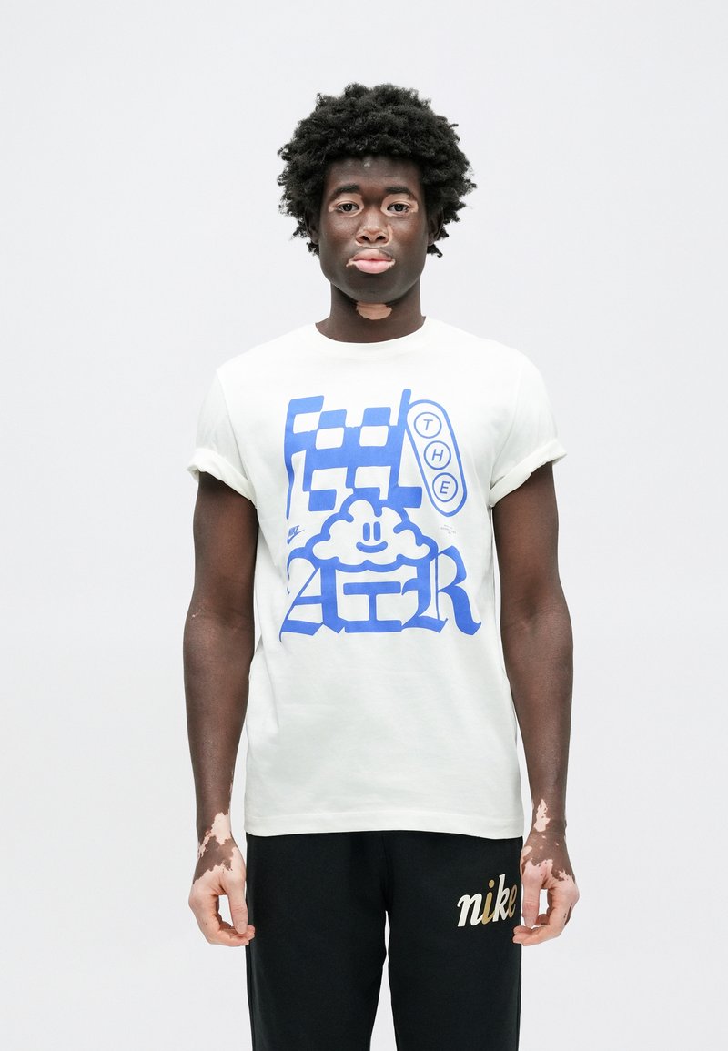 T-shirt blanc à manches courtes avec texte graphique bleu et design de nuage. Porté avec un pantalon de survêtement noir arborant un logo "Nike" doré.