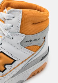 Basket haute New Balance blanche et orange montrant un col de cheville rembourré, des lacets, le logo et une semelle texturée.