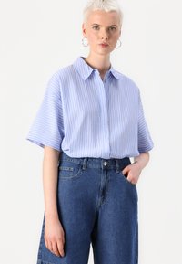 Svetlo modra kratka srajca s kratkimi rokavi in belimi navpičnimi črtami, kombinirana z visokim pasom modrimi jeans hlačami. Gladka tekstura, sproščen kroj.