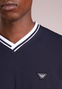 Námořnický polo tričko s výstřihem do V, s bílým pruhovaným límcem a malým vyšitým logem na hrudi. Hladká textura.