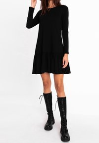 Schwarzes Langarmkleid mit einem gestuften Rüschensaum. Trägt dazu kniehohe schwarze Schnürstiefel. Weicher Stoff, schlichte Silhouette und ohne Muster.