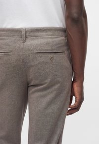 Vue de dos d'une personne portant un pantalon de costume gris avec des poches arrière boutonnées et une chemise blanche, le bras détendu le long du corps.
