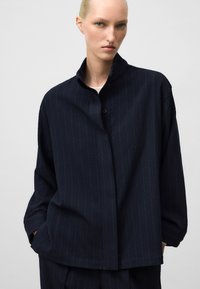 Camicia blu navy con motivo testurizzato, colletto alla mandarina e vestibilità ampia. La camicia presenta righe verticali e tasche laterali.