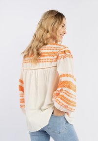 Let bluse lavet af letvægtscreme med orange geometrisk broderi, puffede ærmer og en plisseret rygdesign, sat sammen med denim jeans.