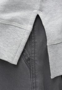 Ύφασμα γκρι φούτερ με ριμπ τελείωμα, επικαλυπτόμενο μερικώς από γκρι παντελόνι. Διαθέτει πλάγια σχισμή και ορατές λεπτομέρειες ραφής.