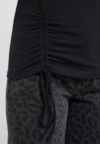 Czarny marszczony top ze ściągaczem u dołu, noszony na szarych spodniach w panterkę. Widok z bliska ukazuje fakturę i szczegóły.