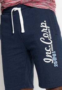 Pantaloni corti in cotone blu con cordino bianco, caratterizzati da un grande testo stampato sulla gamba sinistra e una finitura in tessuto texturizzato.