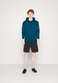 Sweat à capuche teal avec poche kangourou et corde de serrage, associé à un short cargo noir. Le modèle porte des chaussures noires à enfiler. Design simple, sans motifs.
