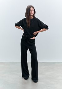 Kvinde med langt hår iført løs sort sweatshirt og brede sorte bukser, der står på grå gulv mod en tom hvid væg.