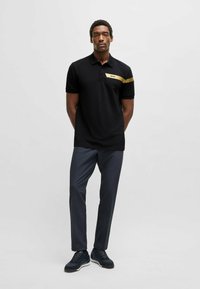 Sort polo skjorte med korte ærmer, med en gylden vandret stribe og logo. Sammen med mørkegrå bukser og sorte sneakers.