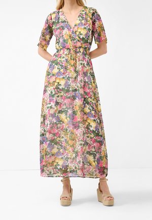Femme portant une robe maxi fleurie avec des fleurs roses, jaunes et violettes et des sandales compensées beiges, debout les mains dans le dos.