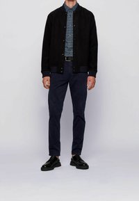 Zwarte gestructureerde bomberjack, blauwe gemusterde shirt, marineblauwe broek en zwarte schoenen met dikke zolen. Knopdetails op de jas, ribgebreide manchetten.