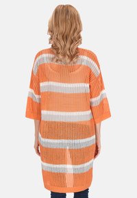 Narancsszínű, kötött takaró, fehér vízszintes csíkokkal, háromnegyedes ujjal, laza fazonnal és textúrált, nyitott szövésű mintával.