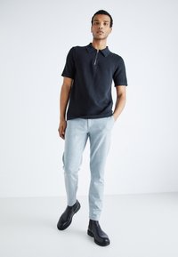 Polo negro de manga corta con cuello con cremallera, combinado con pantalones tapered en gris claro y botas negras de tobillo. Diseño minimalista y textura suave.