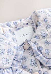 Veste blanche matelassée avec imprimé floral bleu clair, col à carreaux volanté et fermeture par boutons-pression, étiquetée NEXT à l’intérieur de l’encolure.