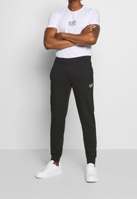 Man met gekruiste armen, gekleed in een wit T-shirt met EA7-logo, zwarte joggers met logo en witte sneakers, staand tegen een effen achtergrond.