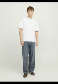 T-shirt bianco in cotone a maniche corte, abbinato a jeans denim blu a vestibilità ampia e sneakers marroni con dettagli bianchi.