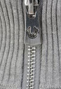 Gros plan sur un tirette de fermeture éclair en métal argenté sur un tissu en maille côtelée gris avec la fermeture éclair partiellement fermée.
