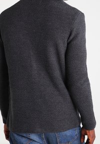 Suéter texturizado gris oscuro con un sutil patrón tejido. Presenta mangas largas y un dobladillo redondeado, combinado con jeans de mezclilla azules.