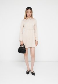 Žena oblečená v svetlo béžových dlhých rukávoch so zapínaním na gombíky, drží malú čiernu kabelku a má na sebe čierne špicaté baleríny.