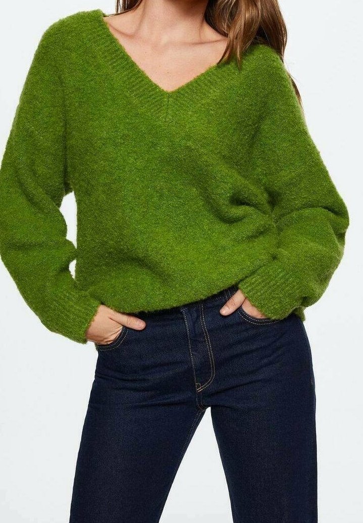 Pull vert en tricot avec col en V, coupe ample, manches longues et poignets côtelés. Porté avec un jean bleu foncé. Texture douce.