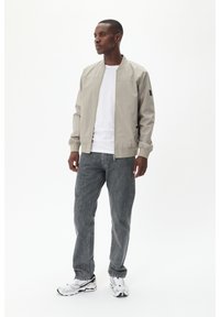 Hellgraue Bomberjacke mit rundem Kragen, Reißverschluss und seitlichen Taschen, kombiniert mit dunkelgrauen Jeans und weißen Sneakers.