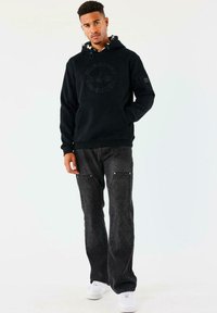 Hombre de pie con las manos en el bolsillo de una sudadera negra, vistiendo jeans negros con parches texturizados y zapatillas blancas sobre un fondo blanco liso.