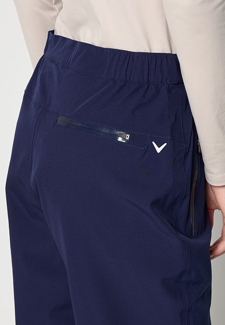 Navyblaue Hose mit elastischem Bund, Reißverschluss am hinteren Taschen, seitlichem Reißverschluss und weißem Häkchen-Logo an der Hüfte, getragen mit beigem Oberteil.