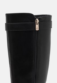 Black faux leather knee-high boot with side zipper, flat sole, and a strap at the top. Smooth texture and sleek design.