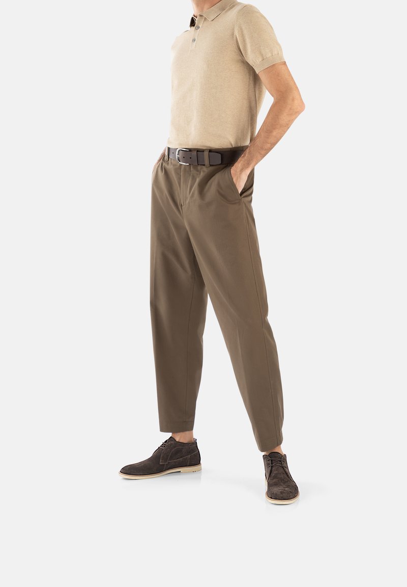 Polo shirt beige à manches courtes, pantalon vert olive, ceinture noire et chaussures en daim marron foncé avec semelles crème. Le mannequin se tient dans une pose neutre.