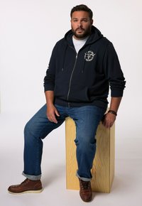 Hoodie zippé bleu marine avec poche avant et logo, assorti à un jean bleu et des bottes en cuir à lacets marron, assis sur un bloc de bois.