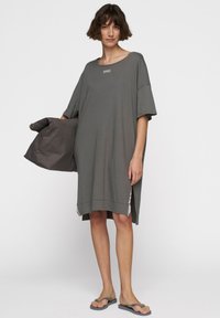 Szara, oversize'owa sukienka t-shirtowa z krótkimi rękawami i bocznymi rozcięciami. Posiada prosty design i metkę przy dekolcie. Nosi szarą torbę.