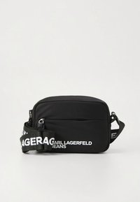 Sort skulderveske med et glatt nylon-yttermateriale, med en frontlomme med glidelås, et trykt logo på stroppen og en justerbar stropp.