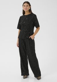 Donna che indossa un top a maniche corte nero con paillettes e pantaloni a palazzo, abbinati a scarpe con tacco a strisce zebrate, in piedi con una mano in tasca.