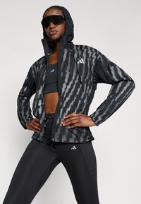 Giacca nera a fantasia con cappuccio, caratterizzata da un design testurizzato in tonalità di grigio. Indossata sopra un top sportivo nero e leggings con accenti di logo.