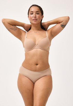 Kvinde med tatoveringer iført beige bh og undertøj med hænderne bag hovedet mod ensfarvet baggrund.