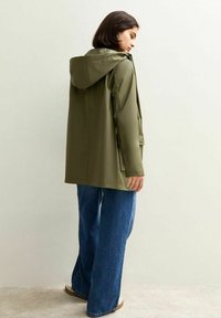 Chaqueta impermeable verde oliva con capucha, de corte recto y con bolsillos laterales, combinada con unos vaqueros de pierna ancha azul.