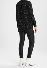 Blusa nera a pois con maniche lunghe, orlo curvo, indossata con leggings neri lucidi e stivaletti bianchi alla caviglia, su uno sfondo semplice.