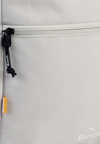 Helle graue Rucksack aus strukturiertem Material, mit einem schwarzen Reißverschluss, einem Zugbändchen und einem weißen Logo an der Basis. Ein kleines orangefarbenes Etikett ist sichtbar.